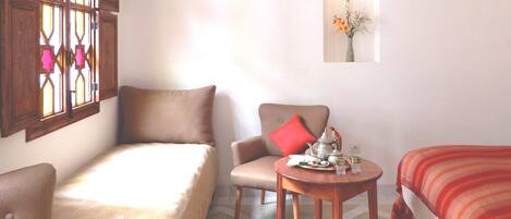 A cozy room with a bed, a chair, a small table with a teapot, and a window with decorative glass.