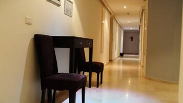 A hallway with two dark-colored chairs and a table, a fire extinguisher on the wall, and a light-colored floor.