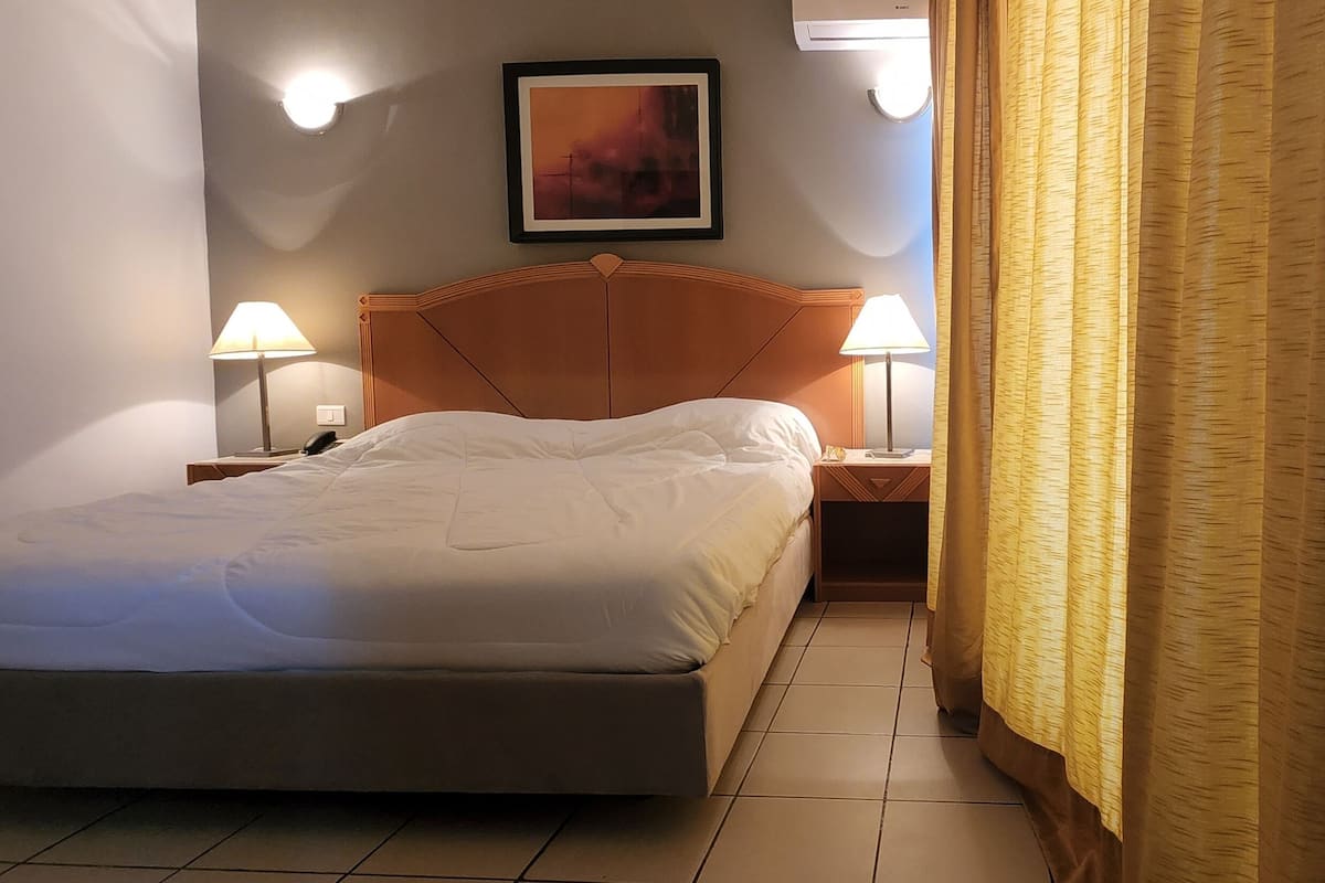 Une chambre d’hôtel avec un lit, deux lampes de chevet, un tableau encadré au mur et un rideau à la fenêtre.