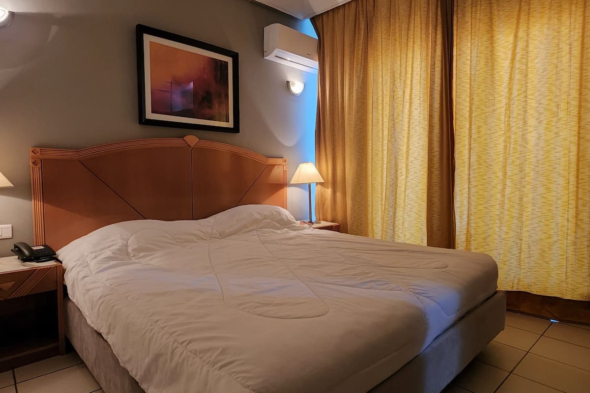 Une chambre d’hôtel avec un lit, deux lampes de chevet, un téléphone, un tableau encadré au mur et une fenêtre avec des rideaux.