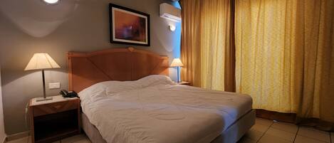 Ein Hotelzimmer mit einem Bett, zwei Nachttischlampen, einem Telefon, einem gerahmten Bild an der Wand und einem Fenster mit Vorhängen.