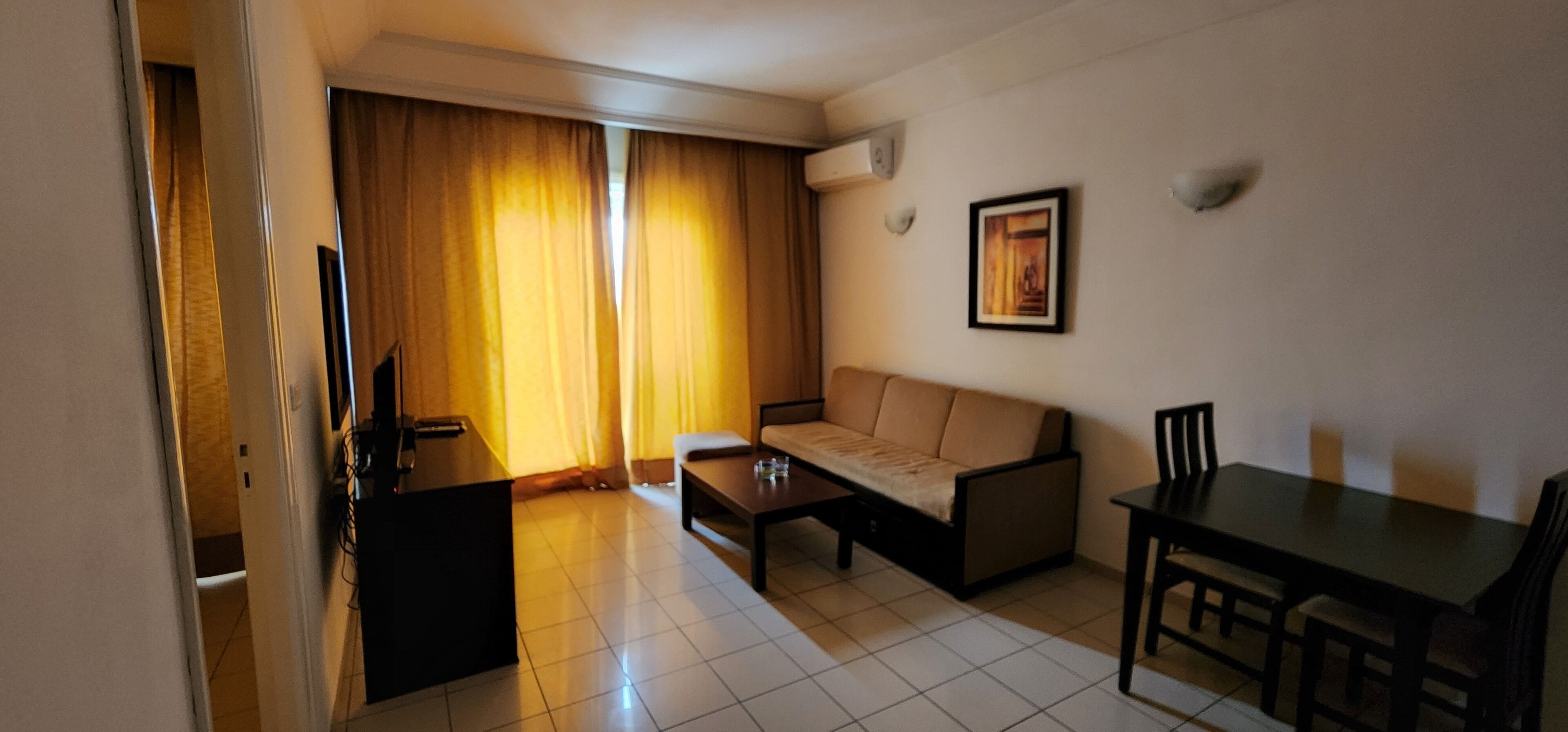Una sala de estar con sofá, mesa de centro y mesa de comedor. Hay un televisor sobre un mueble bajo y un cuadro en la pared.