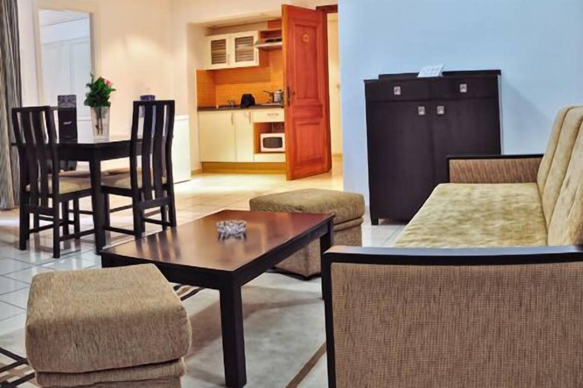 Un salon comprenant un canapé, un pouf et une table à manger avec des chaises. On aperçoit également une cuisine à travers une porte ouverte.