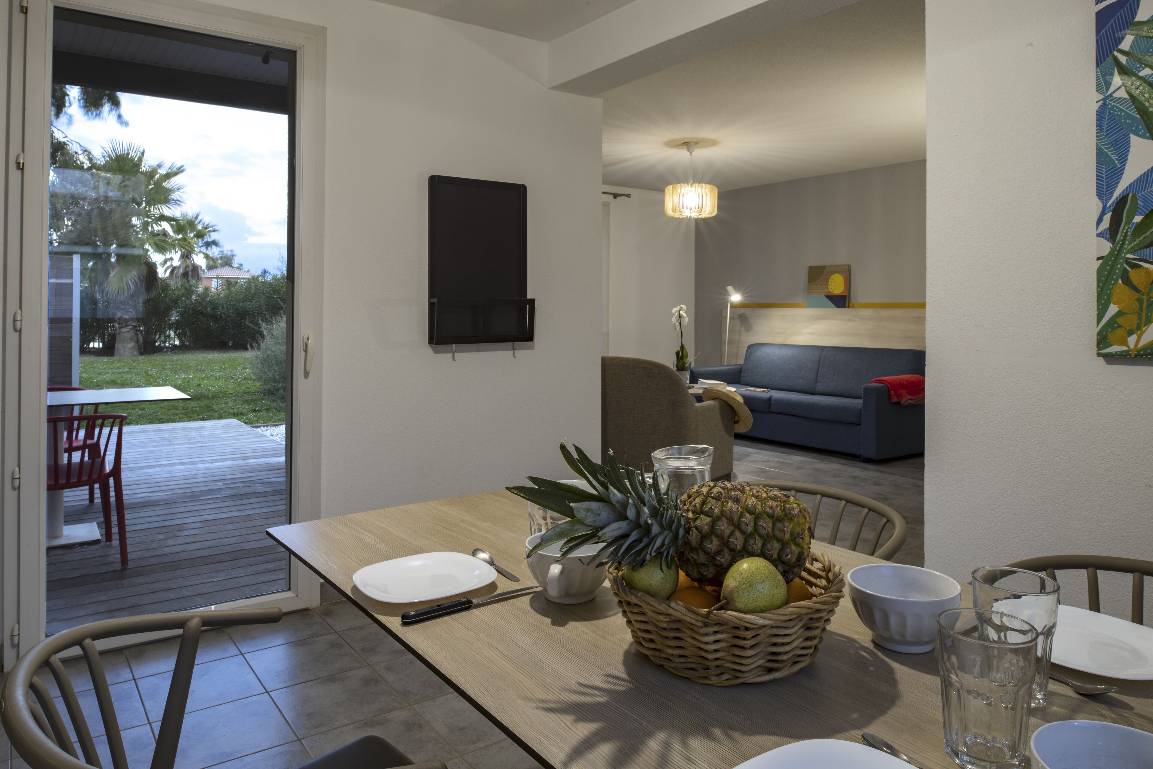 A dining area with a table set for two, a wicker fruit basket, and a view of the outdoor patio.