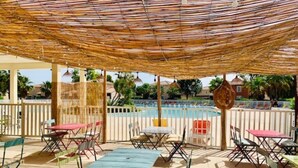 Un patio al aire libre con mesas y sillas, una piscina al fondo y un techo de paja.