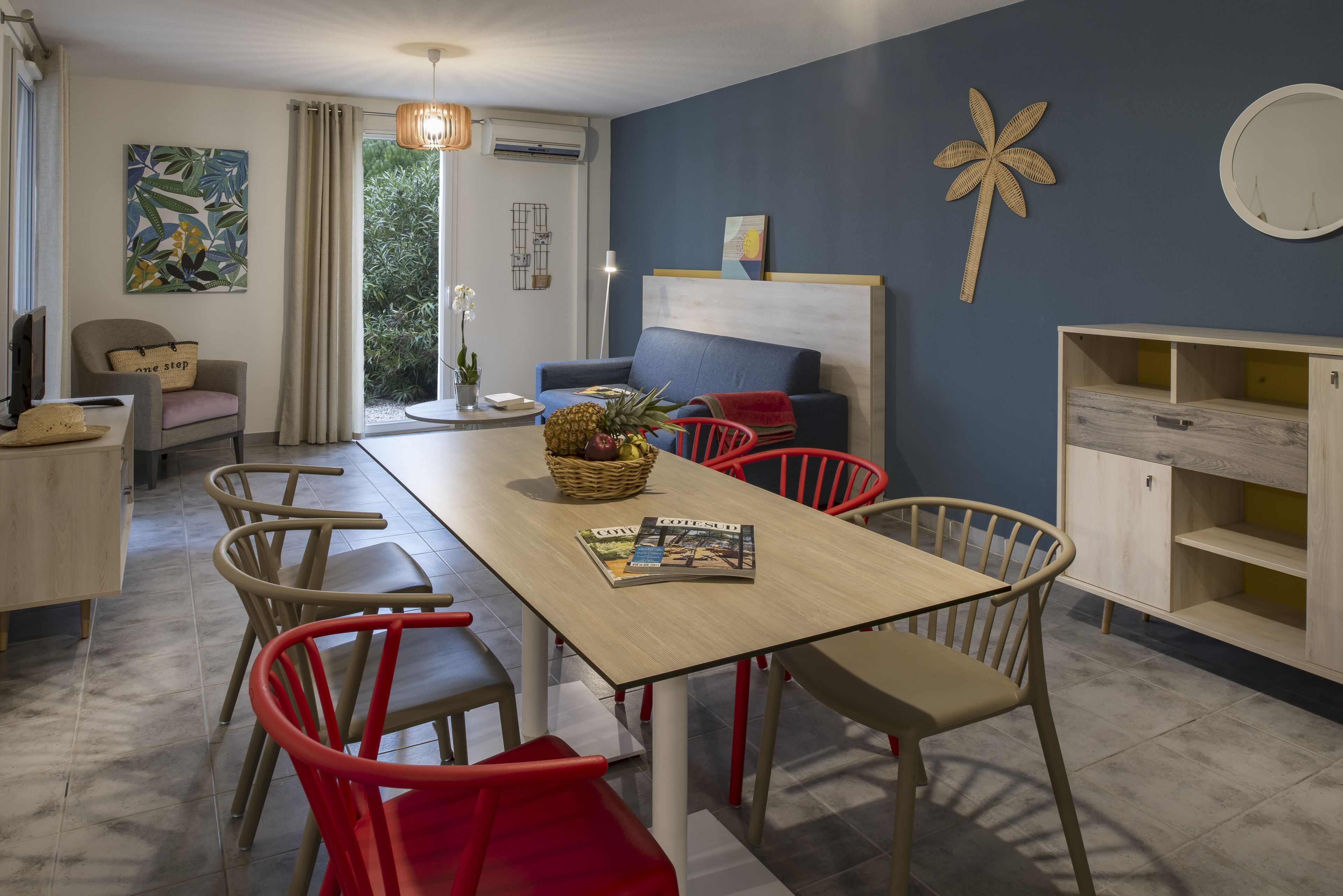 A modern living room with a sofa, dining table, and chairs. There is a window with curtains, a wall art piece, and a small plant on the table.