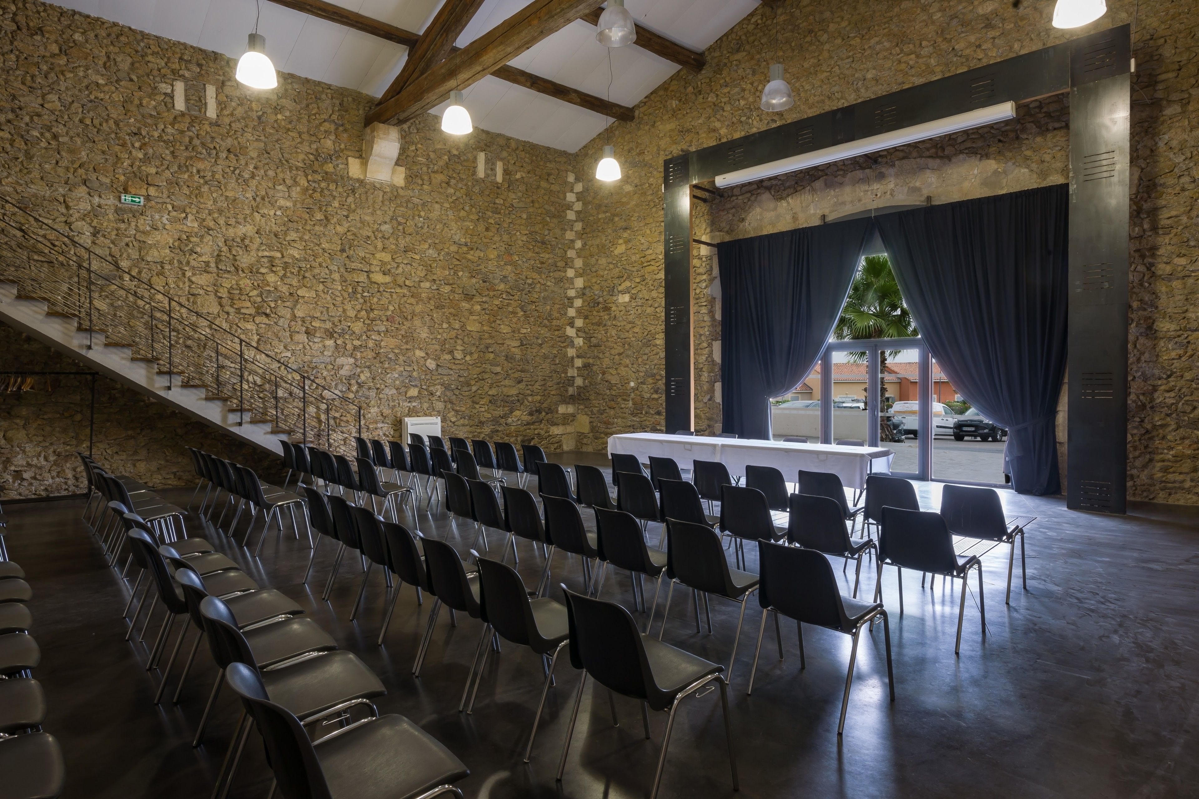 Un salón de conferencias con filas de sillas, una mesa blanca, paredes de piedra y una escalera.