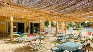 Een terras buiten met rieten dak, meerdere tafels en stoelen, en een zwembad op de achtergrond.