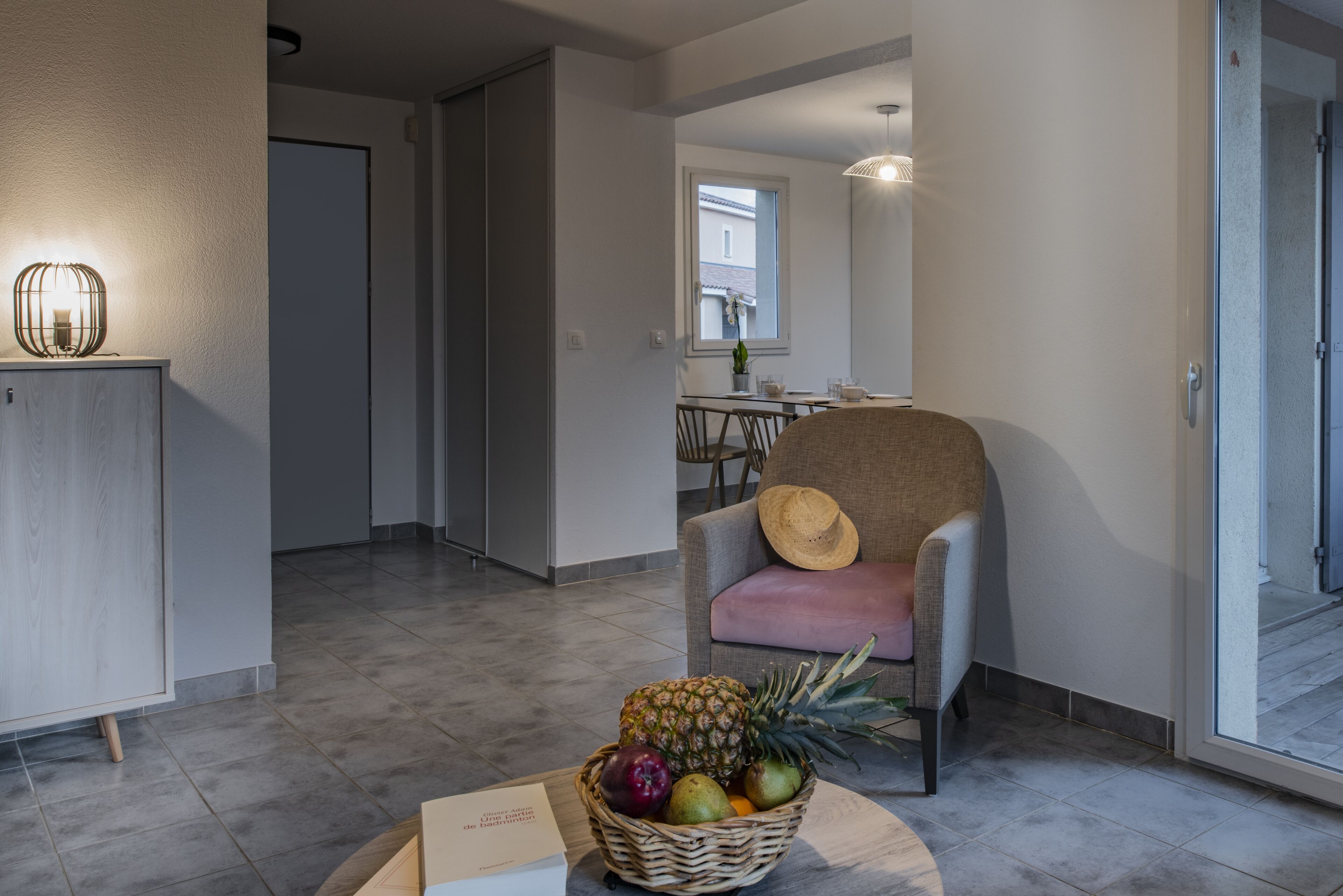 A modern interior with a wicker fruit basket on a table, a chair with a hat, and a dining area in the background.