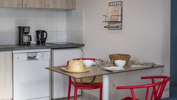 Cocina compacta con cafetera, hervidor y lavavajillas. Hay una mesa de comedor con un sombrero y una cesta.
