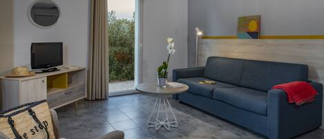 A modern living room with a grey sofa, a round coffee table, a television on a wooden cabinet, and a view of greenery through a sliding door.