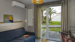 Una sala de estar moderna con un sofá azul, una mesa redonda con libros y un sombrero, una silla roja, y vistas a un jardín a través de una puerta corredera de vidrio.
