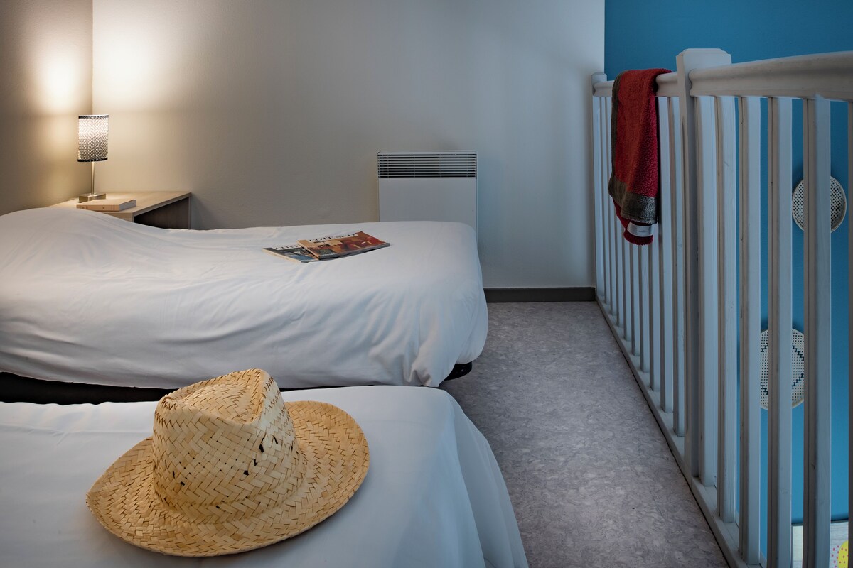 Un dormitorio con dos camas, una mesita de noche con lámpara, un radiador y una cuna con cobija.