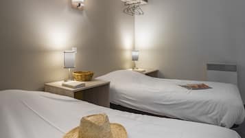 A hotel room with two beds, each with a white bedspread, a nightstand with a lamp, a basket, and a straw hat on one bed.