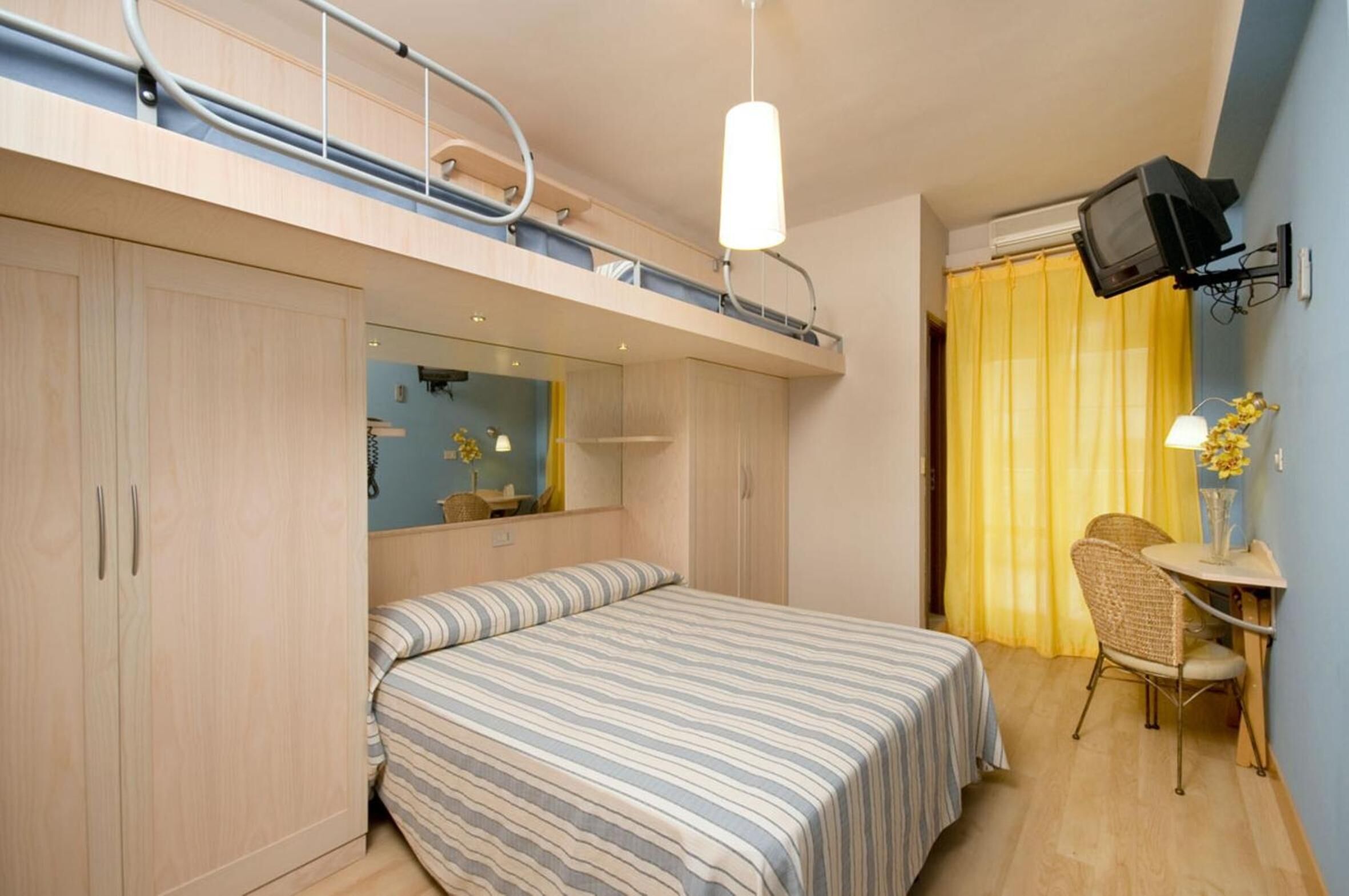 Une chambre d’hôtel compacte comprenant un lit superposé, un petit bureau avec une chaise, un téléviseur fixé au mur et un miroir au-dessus du lit.