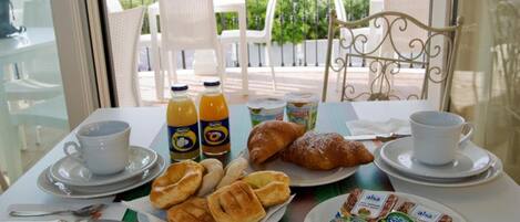 A breakfast table with croissants, pastries, and beverages, set on a balcony with a view of buildings and greenery.