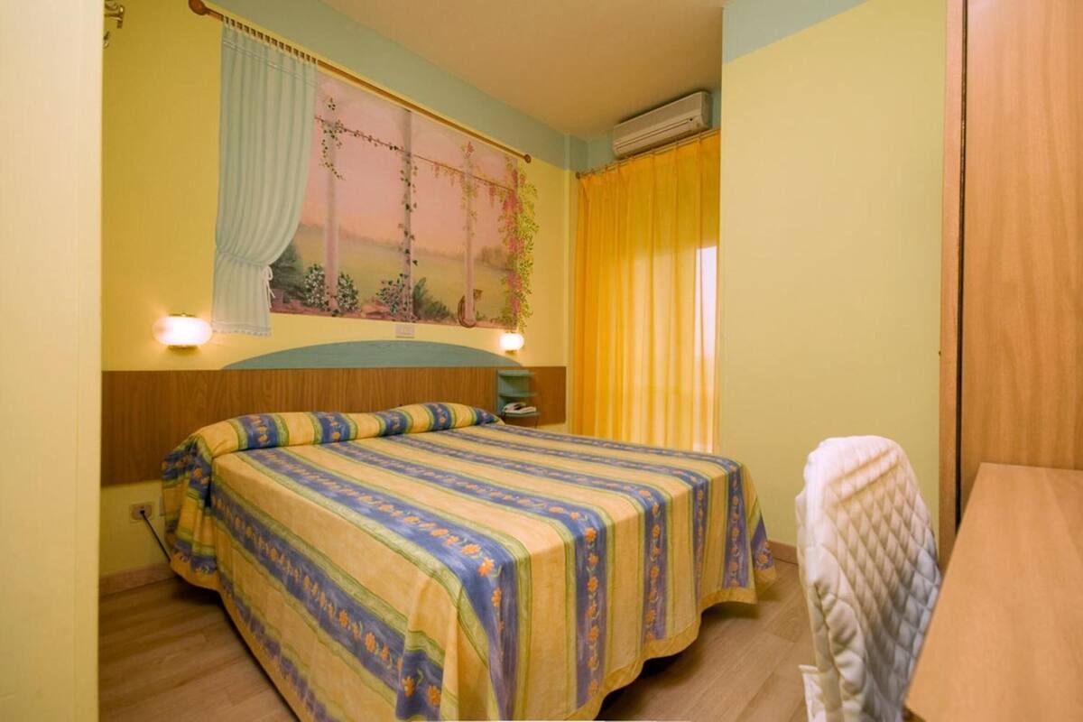 Ein Schlafzimmer mit Bett, Nachttisch, einem Fenster mit Vorhängen und einer Wand-Klimaanlage.