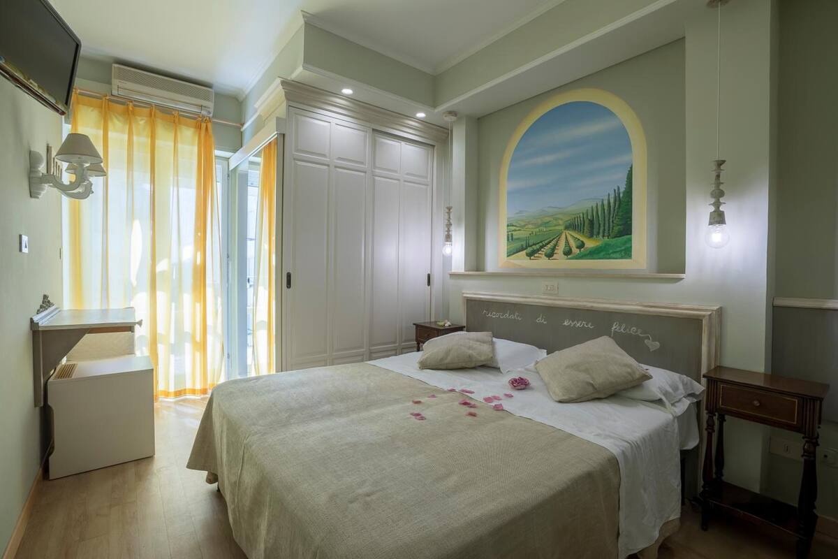 Ein Schlafzimmer mit einem großen Bett, einem Nachttisch, einer Kommode, einem Bild an der Wand und einem Fenster mit Vorhängen.