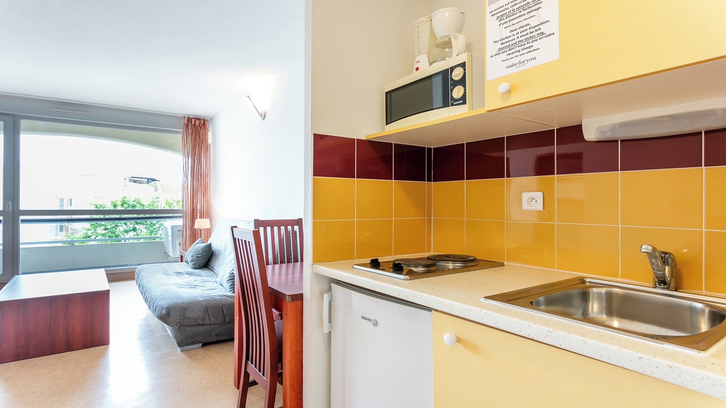 Una cocina compacta equipada con microondas, lavamanos y refrigerador pequeño. Hay un comedor con mesa y sillas, y una ventana con cortinas.