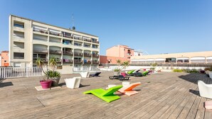 A rooftop deck with lounge chairs, potted plants, and a view of a multi-story building with balconies.