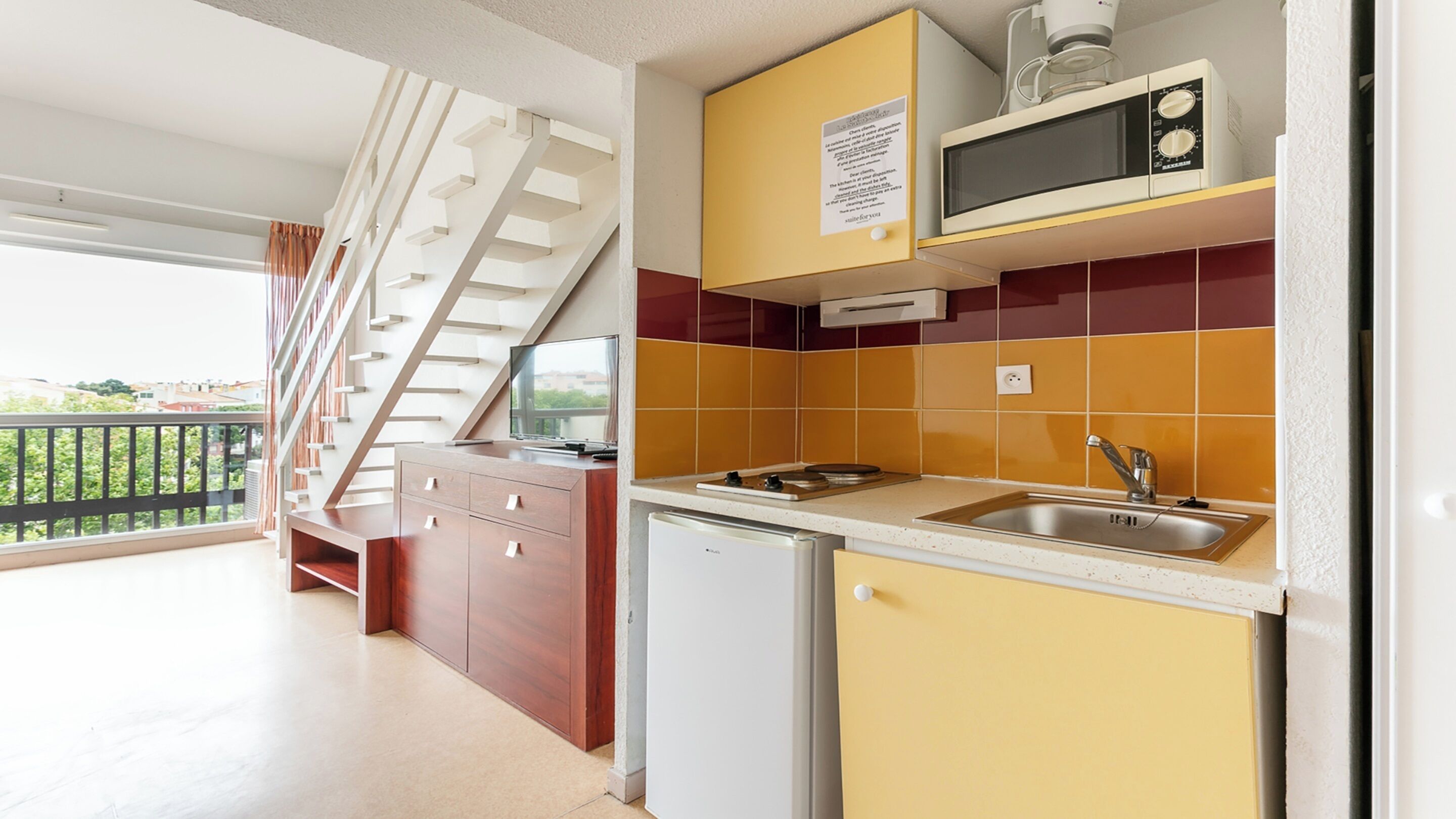 Une cuisine compacte équipée d’un four à micro-ondes, d’un évier et d’un petit réfrigérateur. Un escalier mène à un balcon offrant une vue sur la verdure.