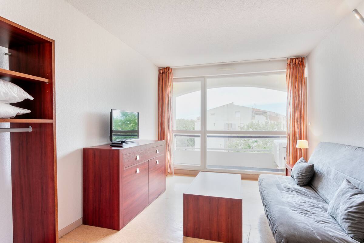 Une chambre d’hôtel moderne avec une armoire en bois, un téléviseur à écran plat, un canapé gris et une grande fenêtre avec des rideaux.