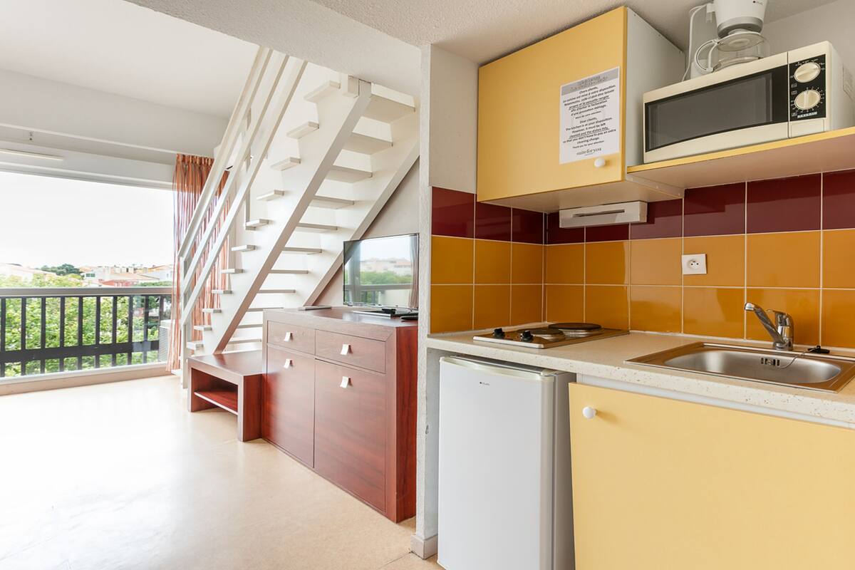 Une cuisine équipée d’un four à micro-ondes, d’un évier et d’un petit réfrigérateur. Un escalier mène à un balcon.
