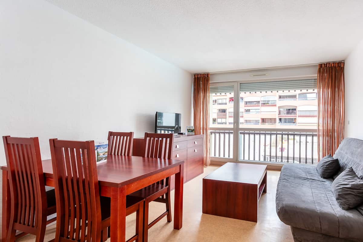 Un salon moderne comprenant un coin repas, un canapé gris, des chaises en bois, un téléviseur et un balcon avec des rideaux.