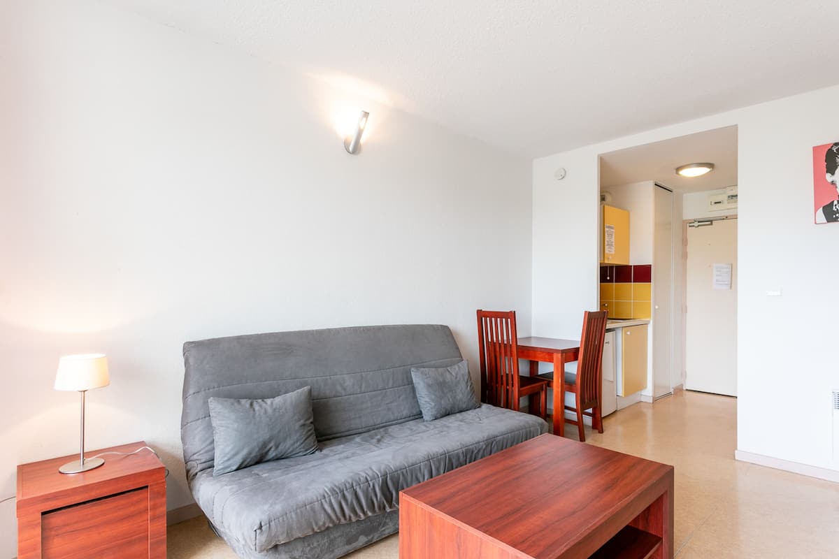 Un salon moderne avec un canapé gris, une table basse en bois, une table d’appoint avec une lampe, et un coin repas avec une table et des chaises en bois.