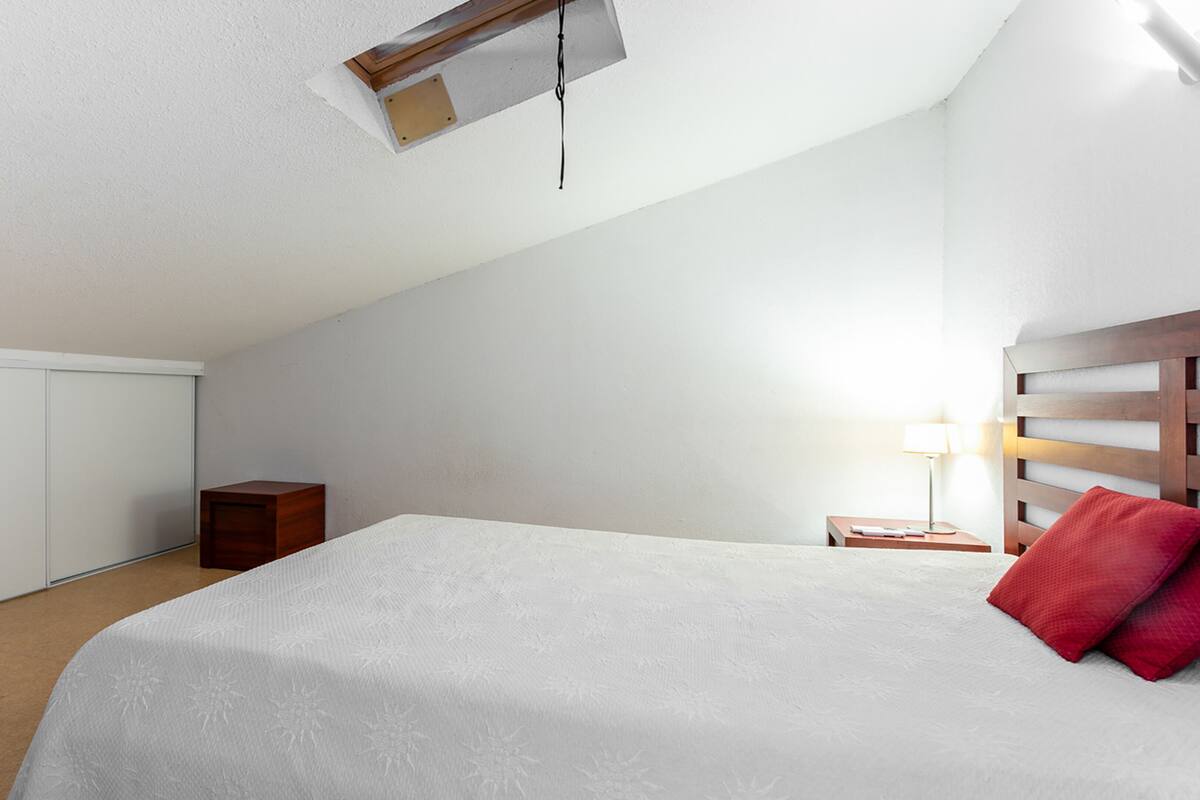 Une chambre avec un plafond en pente, un lit recouvert d’une courtepointe blanche, une tête de lit en bois, une table de chevet avec une lampe et une petite armoire en bois.