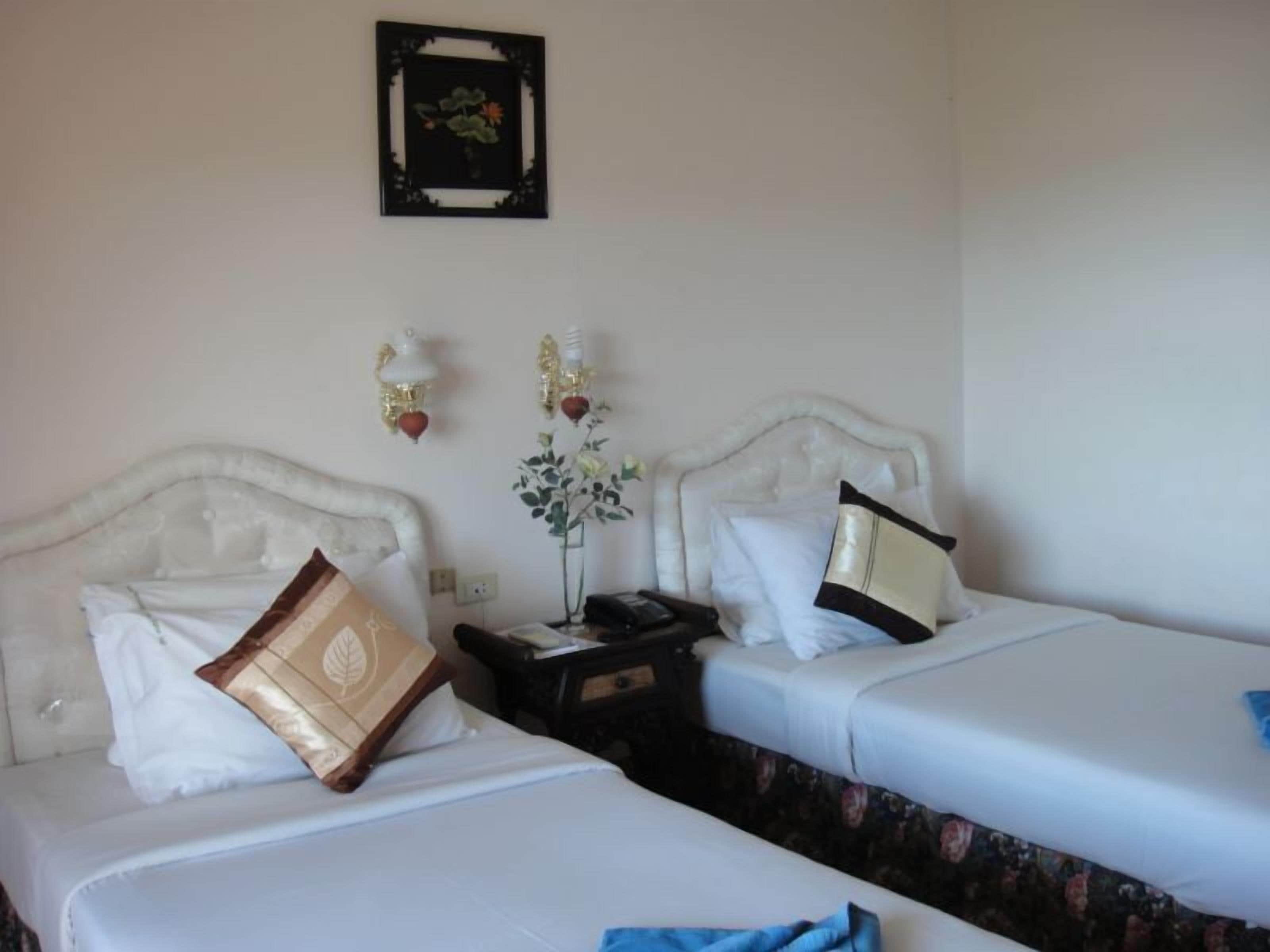A hotel room with two beds, each with a white headboard and a single pillow. There is a nightstand with a lamp and a vase with flowers between the beds.