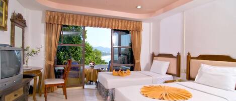 A hotel room with two beds, a television, a dining area with a table and chairs, and a large window offering a view of greenery and mountains.