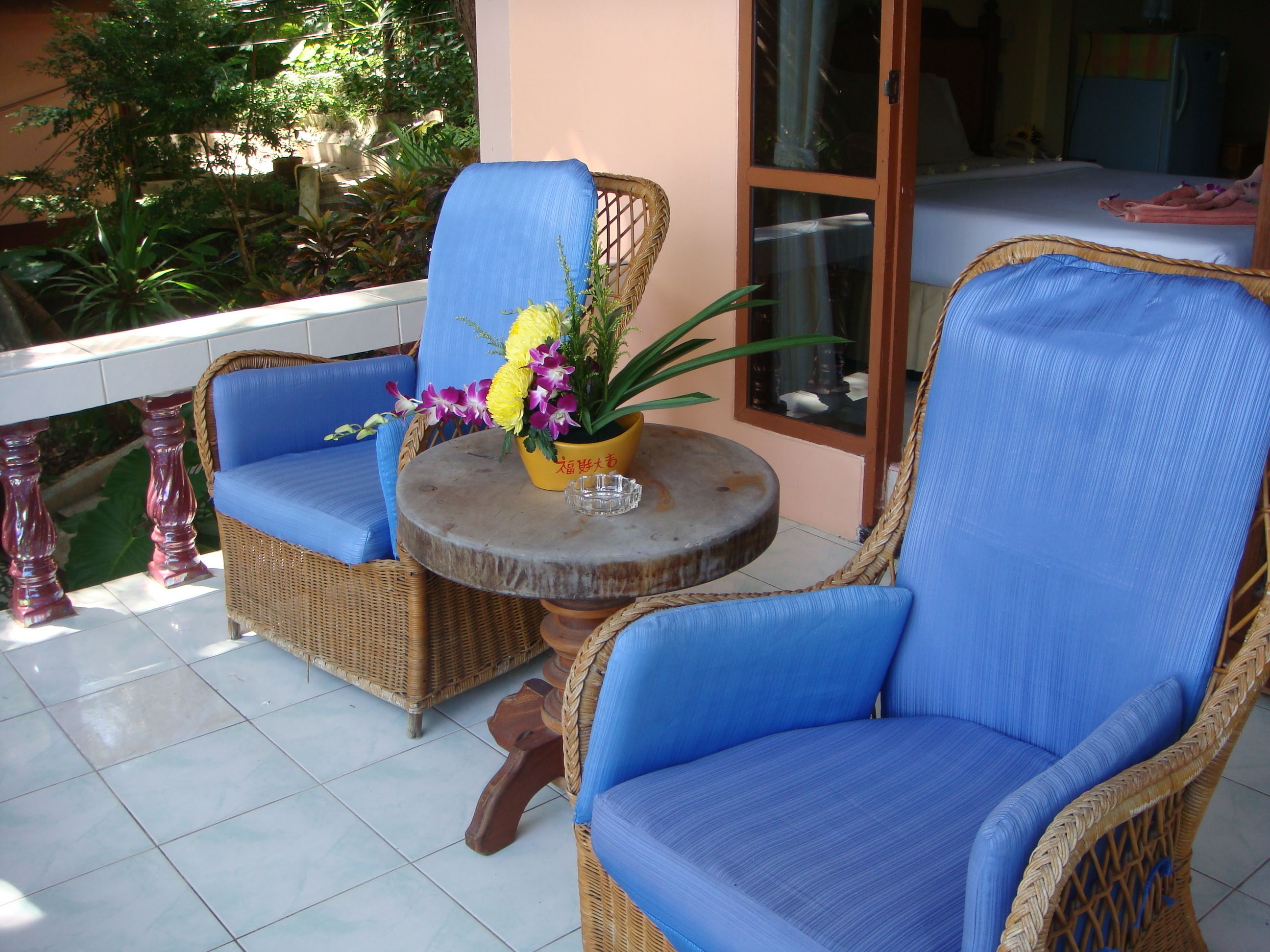 A balcony with wicker chairs and a small table, a potted plant, and a glimpse of a room with a bed and window.