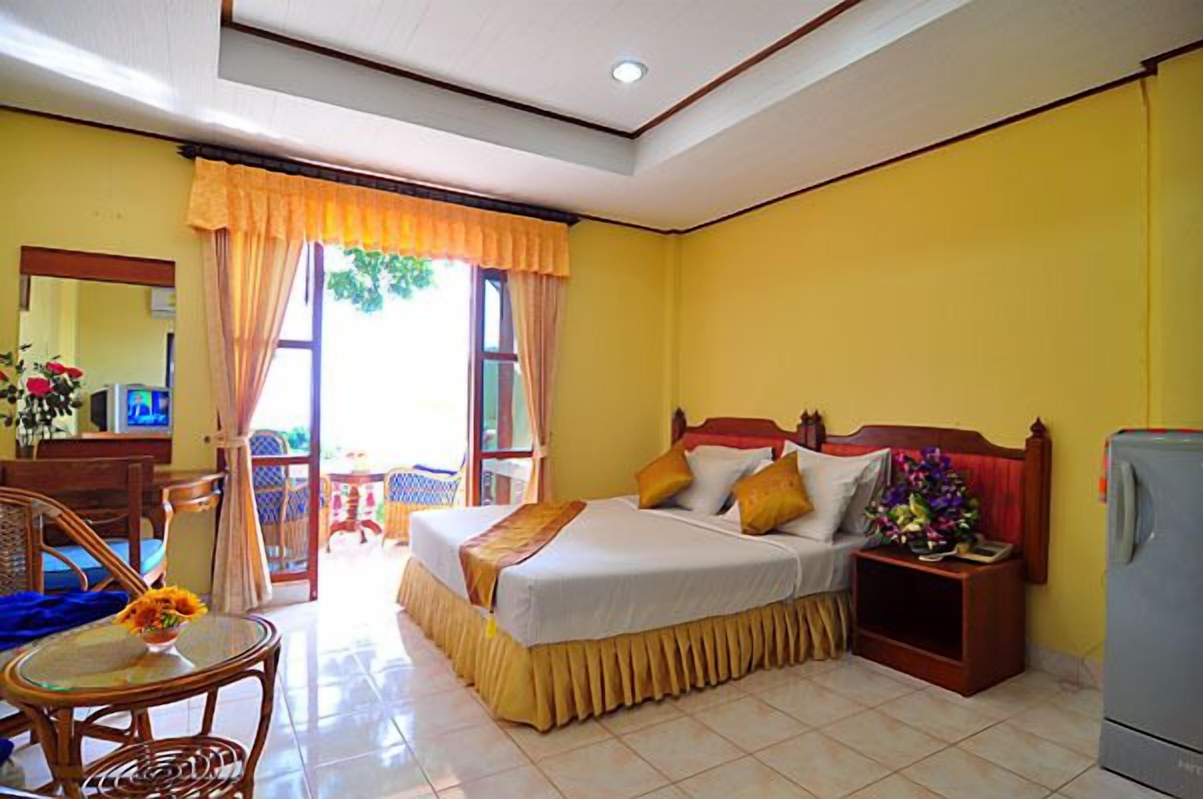 A hotel room with a bed, a small table with a vase, a chair, a nightstand, and a refrigerator.