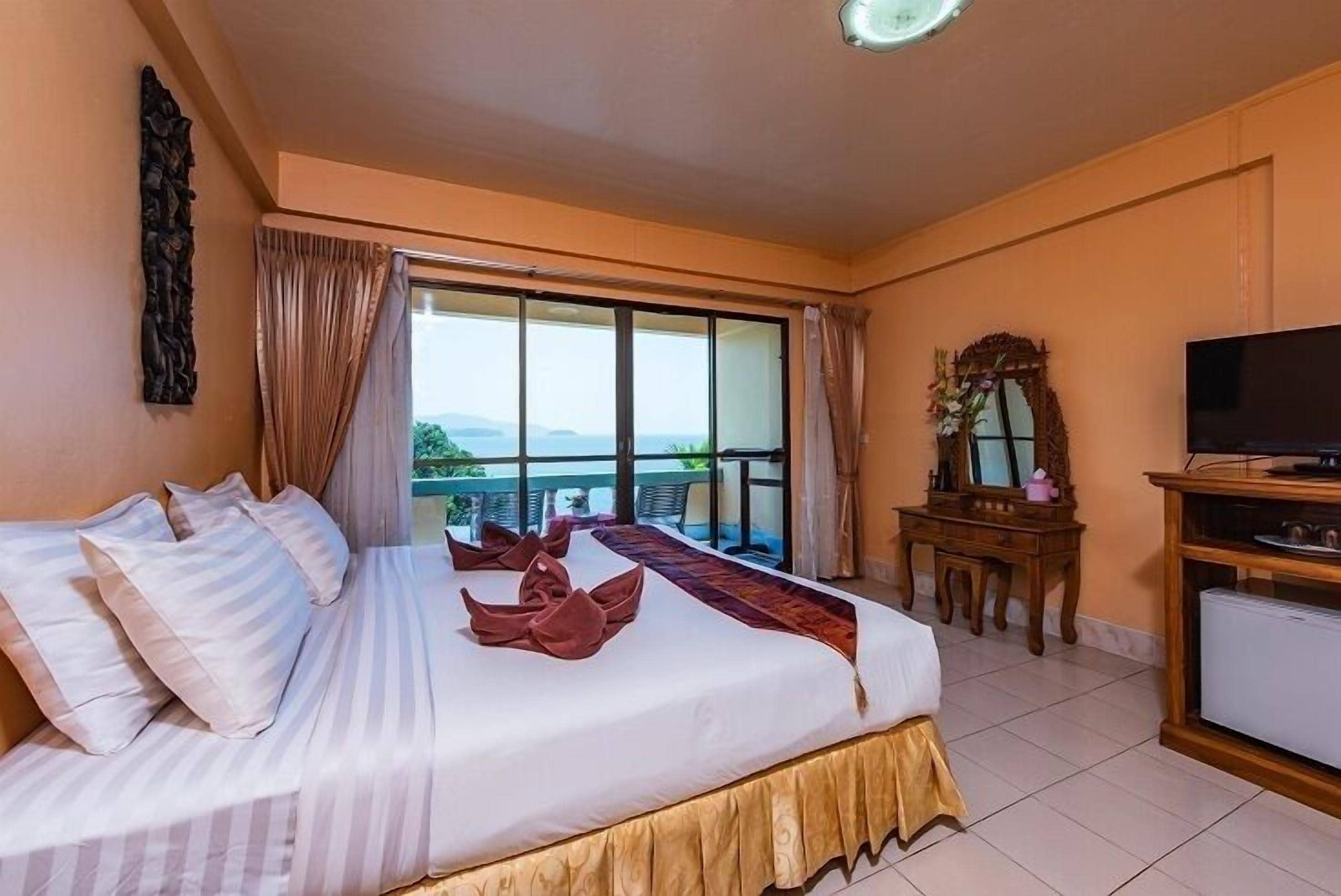 A hotel room with a large bed, a television, a wooden side table, and a view of the outdoors through a large window.