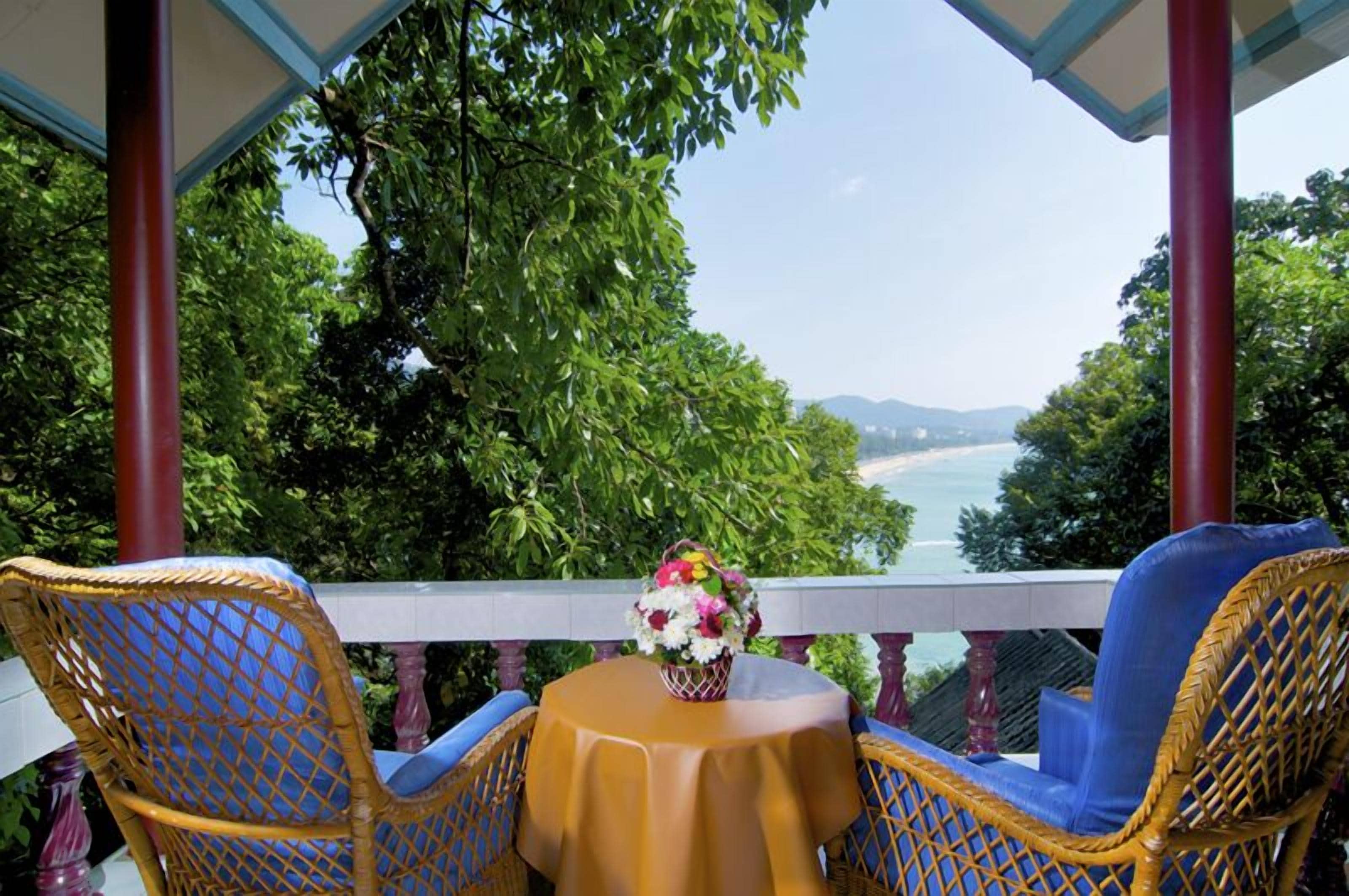 Un balcon agrémenté de fauteuils et d’une table en osier, offrant une vue panoramique sur la mer et la verdure.