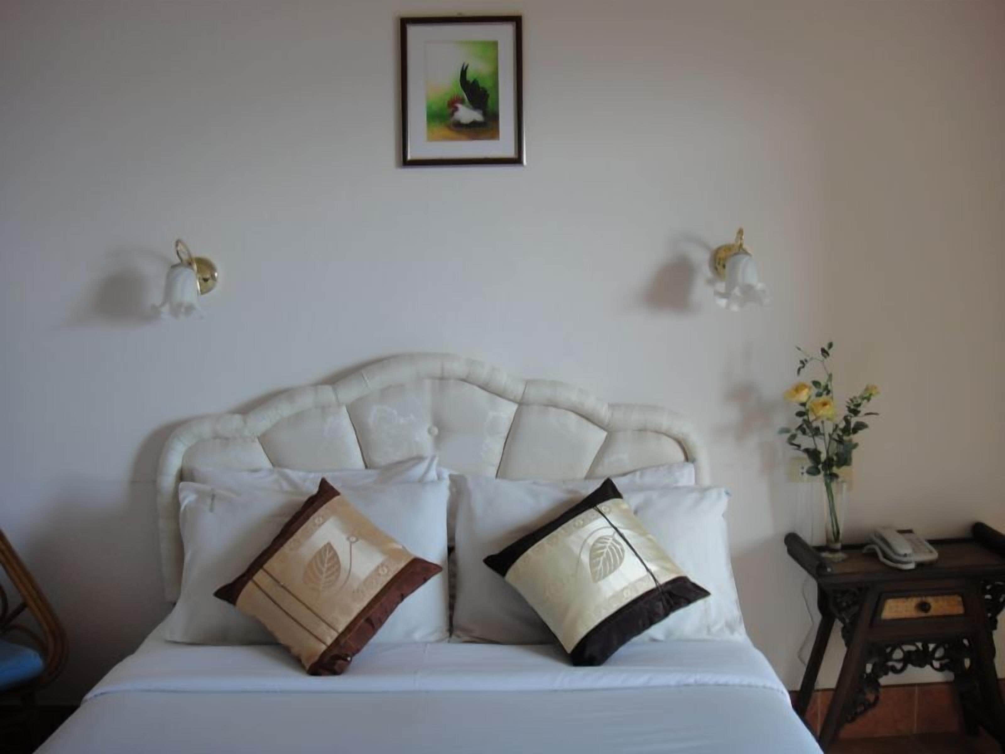 Un lit bien fait, doté d’une tête de lit capitonnée, de deux coussins décoratifs, d’une œuvre d’art encadrée accrochée au mur et d’une petite table sur laquelle est posé un vase de fleurs.