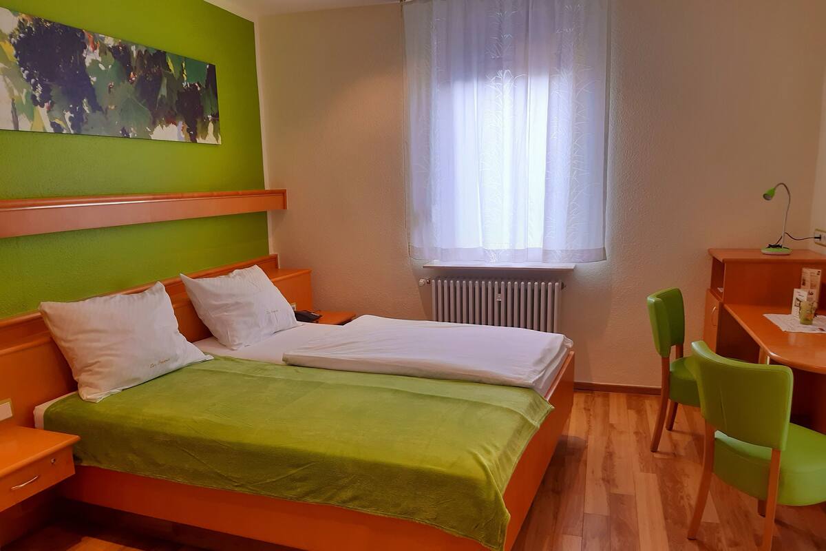 A bedroom with a bed, a desk, a chair, a radiator, and a window with sheer curtains.