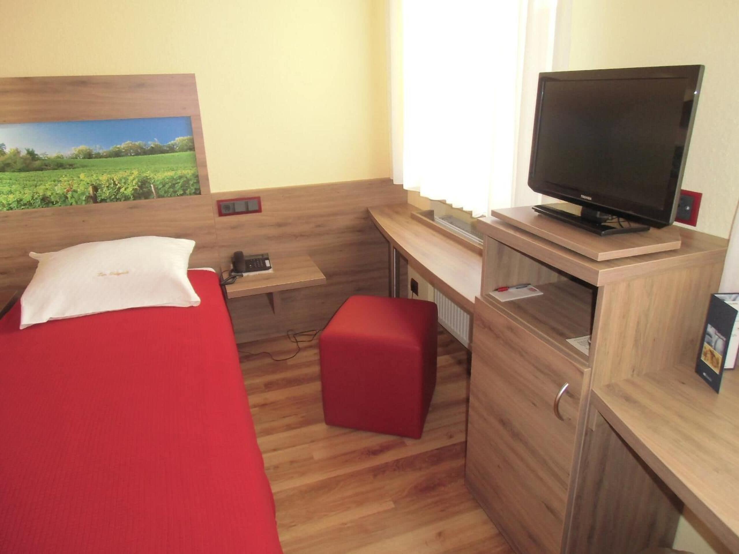 Une chambre d’hôtel compacte, comprenant un lit simple, un bureau avec un ordinateur, une télévision et un petit banc rouge.
