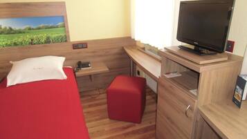 Une chambre d’hôtel compacte, comprenant un lit simple, un bureau avec un ordinateur, une télévision et un petit banc rouge.