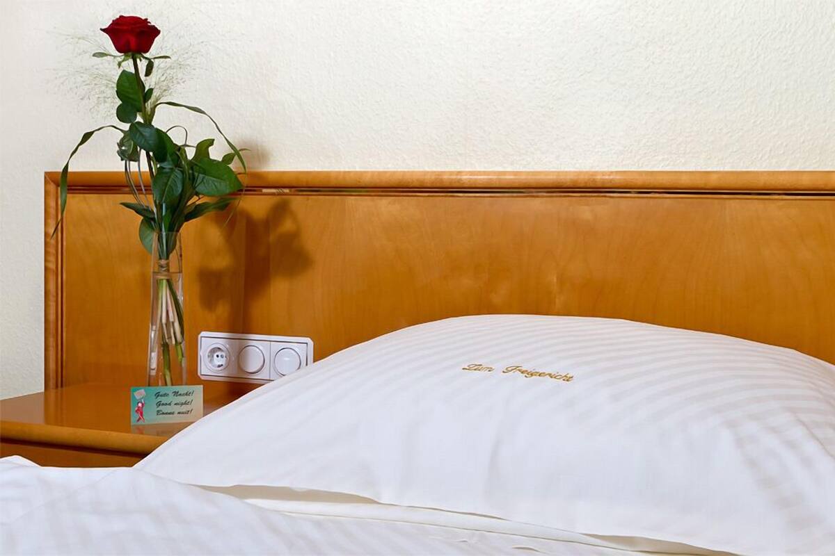 A hotel room with a wooden headboard, a bedside table with a vase of a red rose, and a sign with a message.