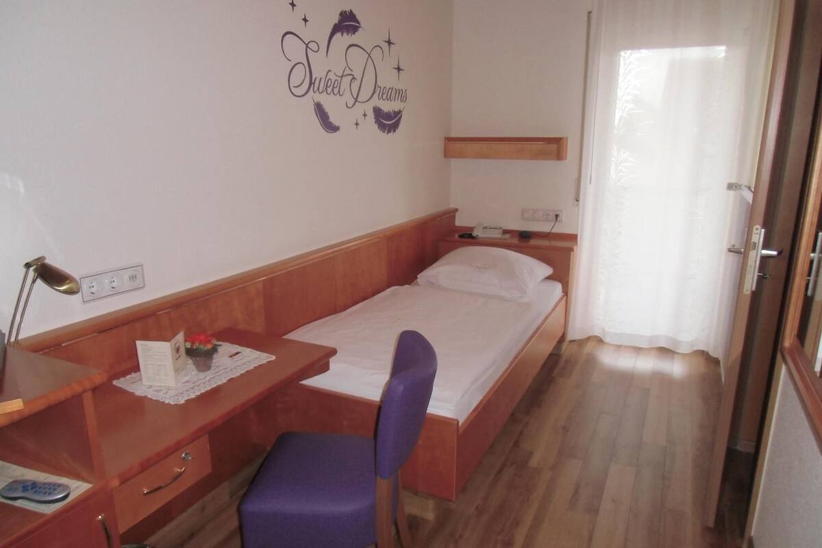A small, neatly arranged bedroom with a single bed, a desk with a chair, and a bedside table.