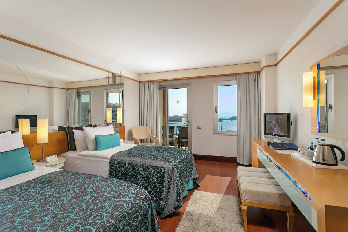 A hotel room with two beds, a desk with a TV, a chair, and a view of the sea.