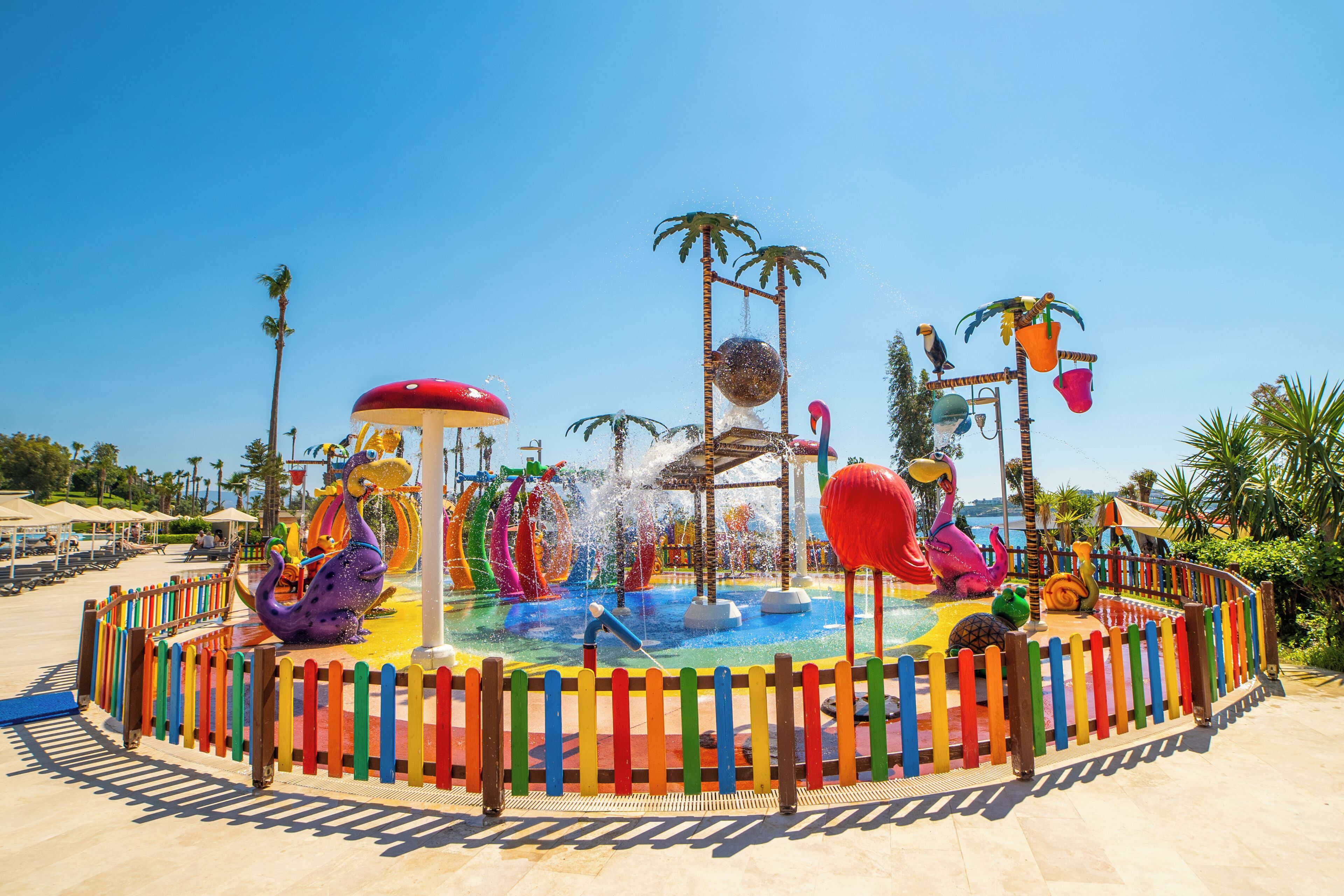 Ein Wasserpark mit einem zentralen Wasserspielplatz, Rutschen und einem farbenfrohen Zaun.