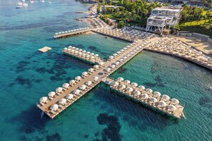 Vista aérea de un resort con varias cabañas sobre el agua conectadas por pasarelas, agua turquesa cristalina y una zona de playa con sombrillas.