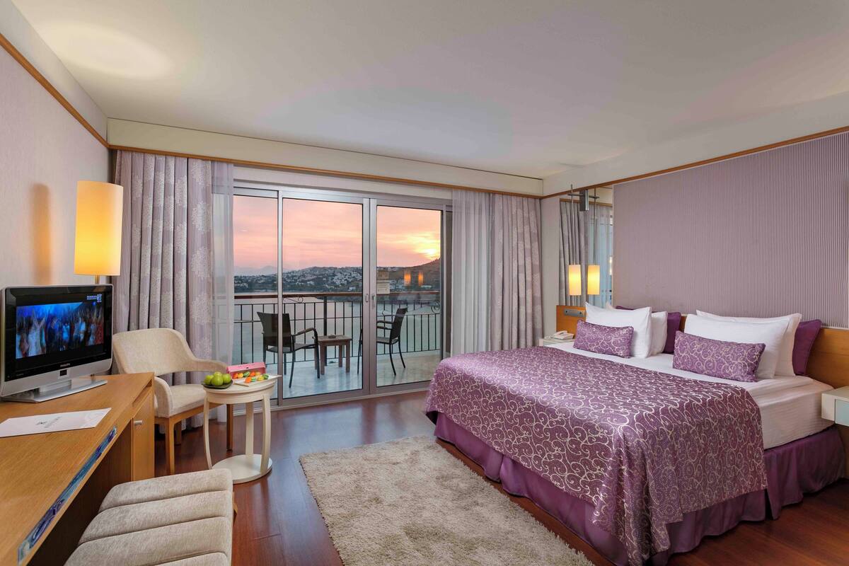 A hotel room with a bed, a desk with a TV, a small table with fruit, and a balcony with a view.