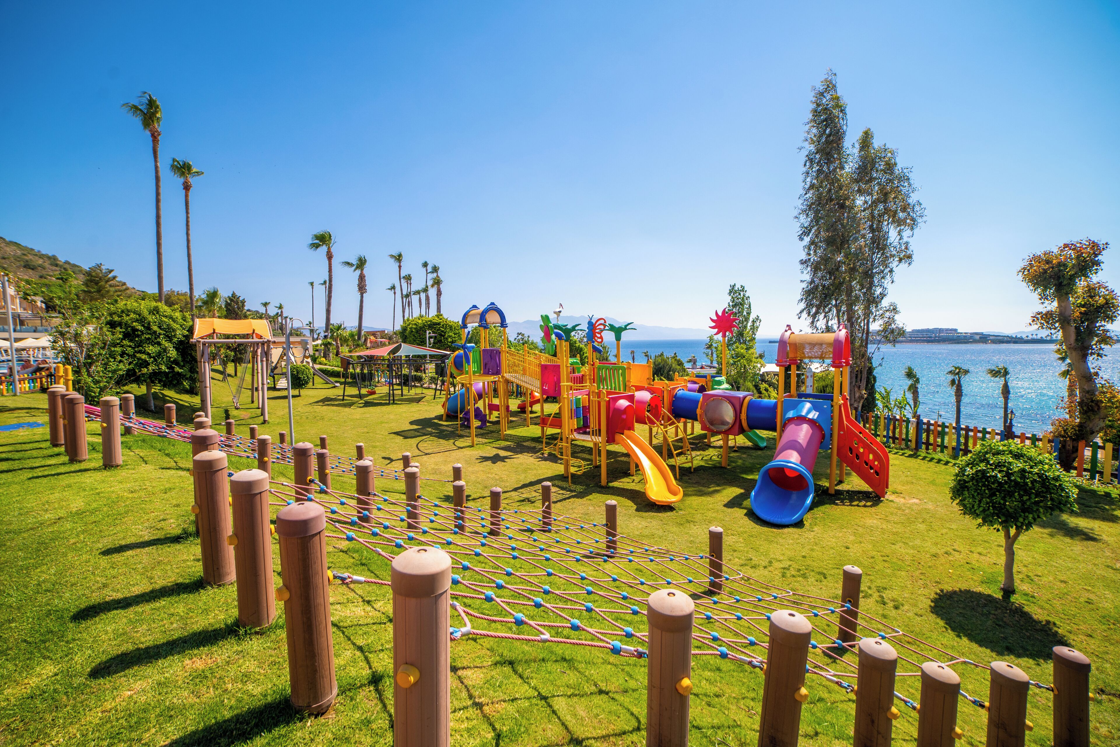 Un parque infantil con diversas estructuras de juego, una red de cuerdas y vistas al mar.