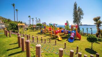 Un parque infantil con diversas estructuras de juego, una red de cuerdas y vistas al mar.