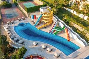 Un área de piscina con tobogán de agua, sillas de descanso y cancha de tenis.