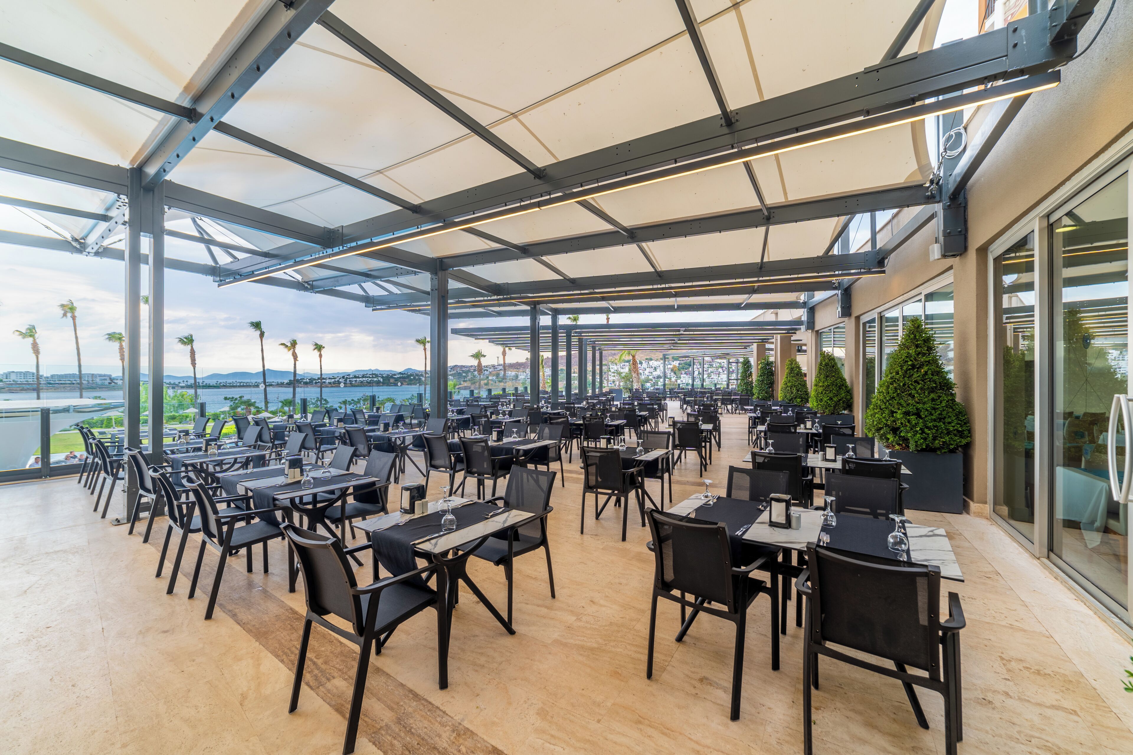 Un área de comedor al aire libre con mesas y sillas, ventanas amplias y vista al agua.